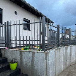 LAKRYS Krystian Lalak - Nowoczesne ogrodzenie metalowe w kolorze grafitowym, zamontowane na betonowym murku oporowym, z widocznymi schodami wejściowymi i rośliną w doniczce.