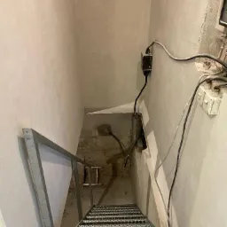 Metalowe schody prowadzące w dół, surowe ściany z widocznymi przewodami elektrycznymi, w oddali ziemia z wystającymi kablami.