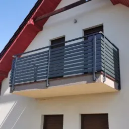 Szary balkon z poziomymi panelami zamontowany na białym budynku z czerwonym dachem i brązowymi roletami w oknach.