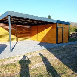 LAKRYS Krystian Lalak - Drewniany carport z szarym dachem i ścianą z poziomych listew, widoczny cień osoby robiącej zdjęcie.