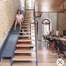 LAKRYS Krystian Lalak - Nowoczesne biuro z nietypowymi schodami: kombinacja drewnianych stopni i metalowej zjeżdżalni. Kobieta zjeżdża na zjeżdżalni. W tle przestrzeń biurowa z biurkami i krzesłami.