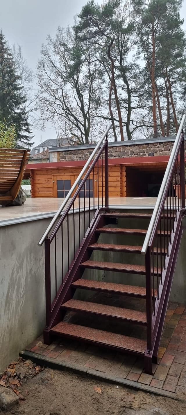 Zewnętrzne schody metalowe z poręczami ze stali nierdzewnej prowadzące do drewnianej altany z kamiennym dachem, widoczne drzewa w tle.