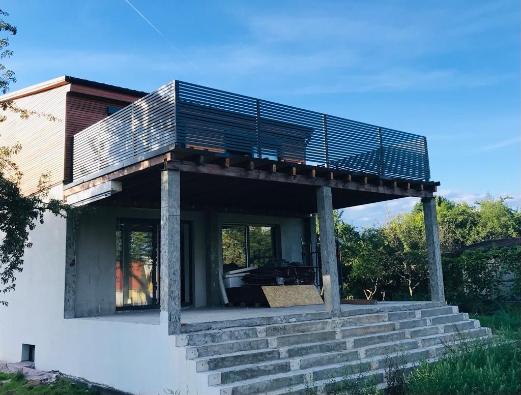 Dom w trakcie budowy z widocznym tarasem z metalową balustradą, surowy beton na schodach i kolumnach, materiały budowlane na tarasie.