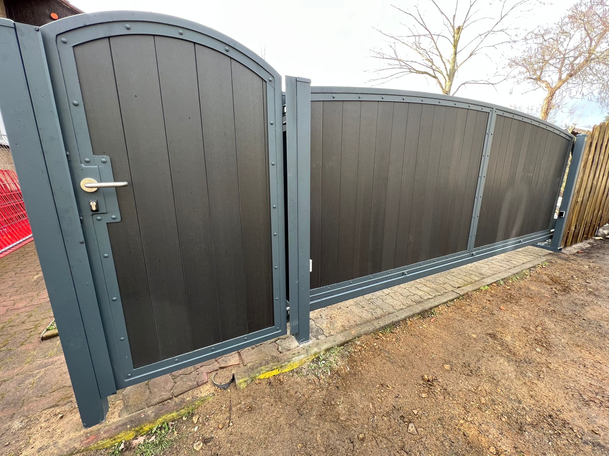 Automatyczna brama wjazdowa z ciemnego drewna kompozytowego z szarymi metalowymi ramami i elementami mocującymi, widoczna z perspektywy ziemi.