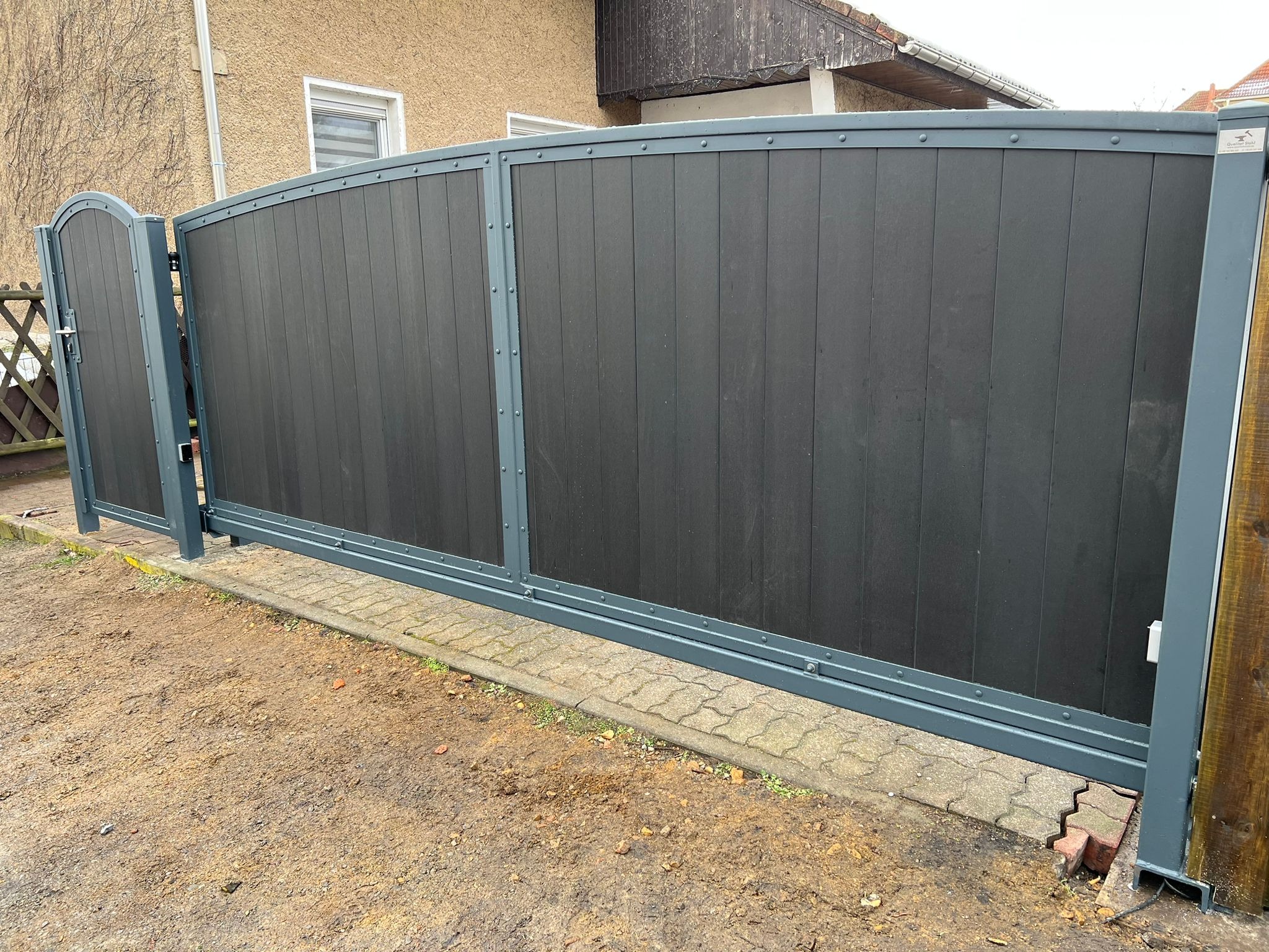 Nowoczesna, przesuwna brama w kolorze grafitowym z szarymi metalowymi elementami konstrukcyjnymi, widoczna z zewnątrz posesji na tle domu.