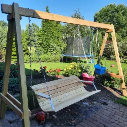 Drewniana huśtawka ogrodowa z ławką i dwoma siedziskami dla dzieci, zawieszona na łańcuchach, na tle zadbanego ogrodu z trampoliną.