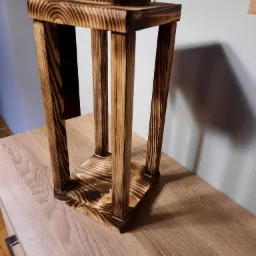 Drewniana, opalana lampionowa konstrukcja z czterema słupkami, widoczna na drewnianym blacie, z gniazdkiem elektrycznym w tle.