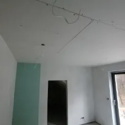 Montaż płyt kartonowo-gipsowych na suficie, widoczne przewody elektryczne i szpachlowane łączenia, surowy stan wykończenia wnętrza.