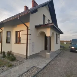 Wykończony dom jednorodzinny z jasną elewacją, brązowymi oknami i dachem, widoczny z boku, z utwardzonym chodnikiem i podjazdem, zielony bus zaparkowany obok.