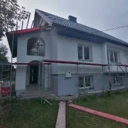 Dwukondygnacyjny dom jednorodzinny w trakcie malowania elewacji, widoczne rusztowania i częściowo pomalowana dolna partia budynku na szaro.