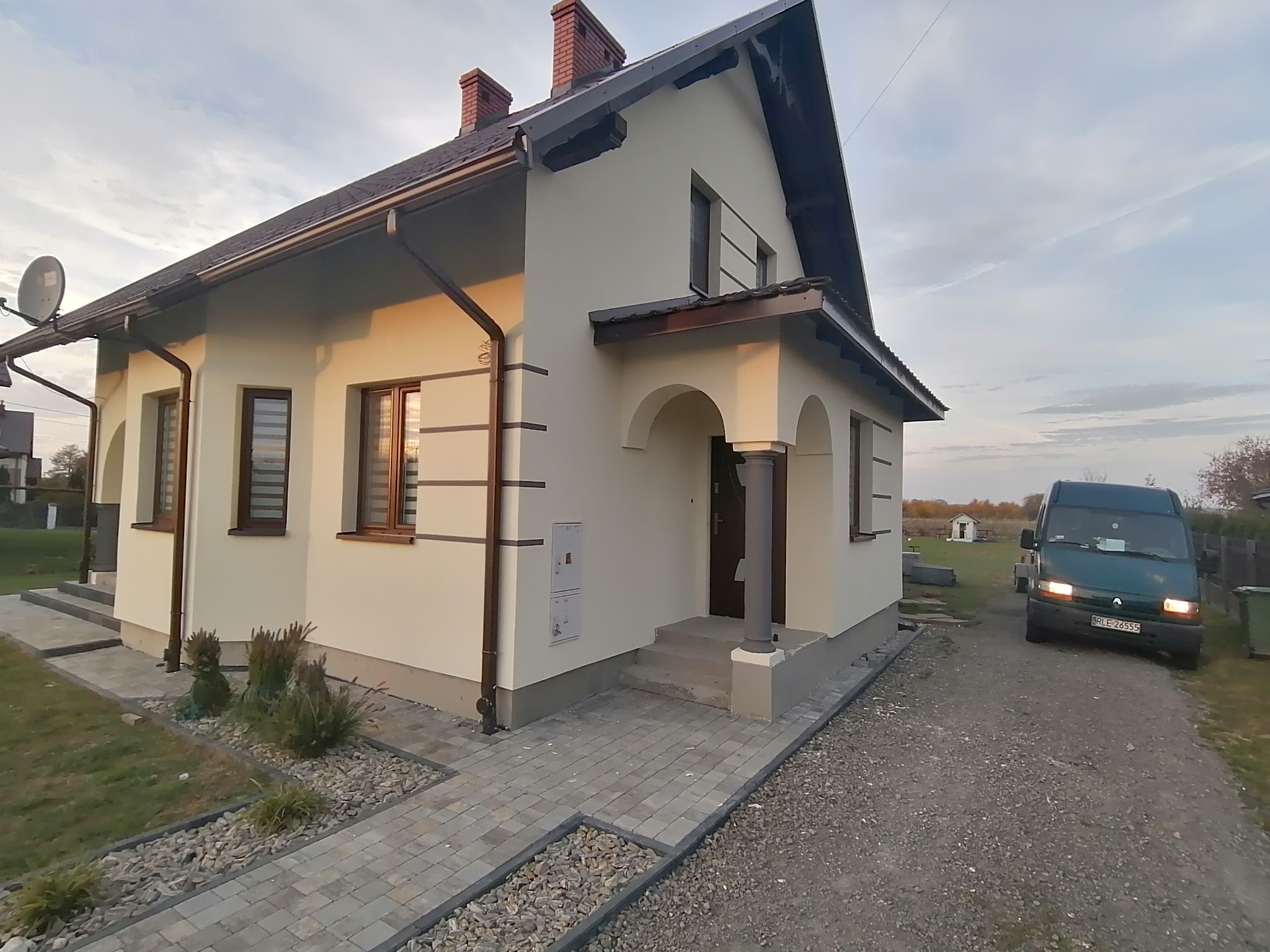 Wykończony dom jednorodzinny z jasną elewacją, brązowymi oknami i dachem, widoczny z boku, z utwardzonym chodnikiem i podjazdem, zielony bus zaparkowany obok.