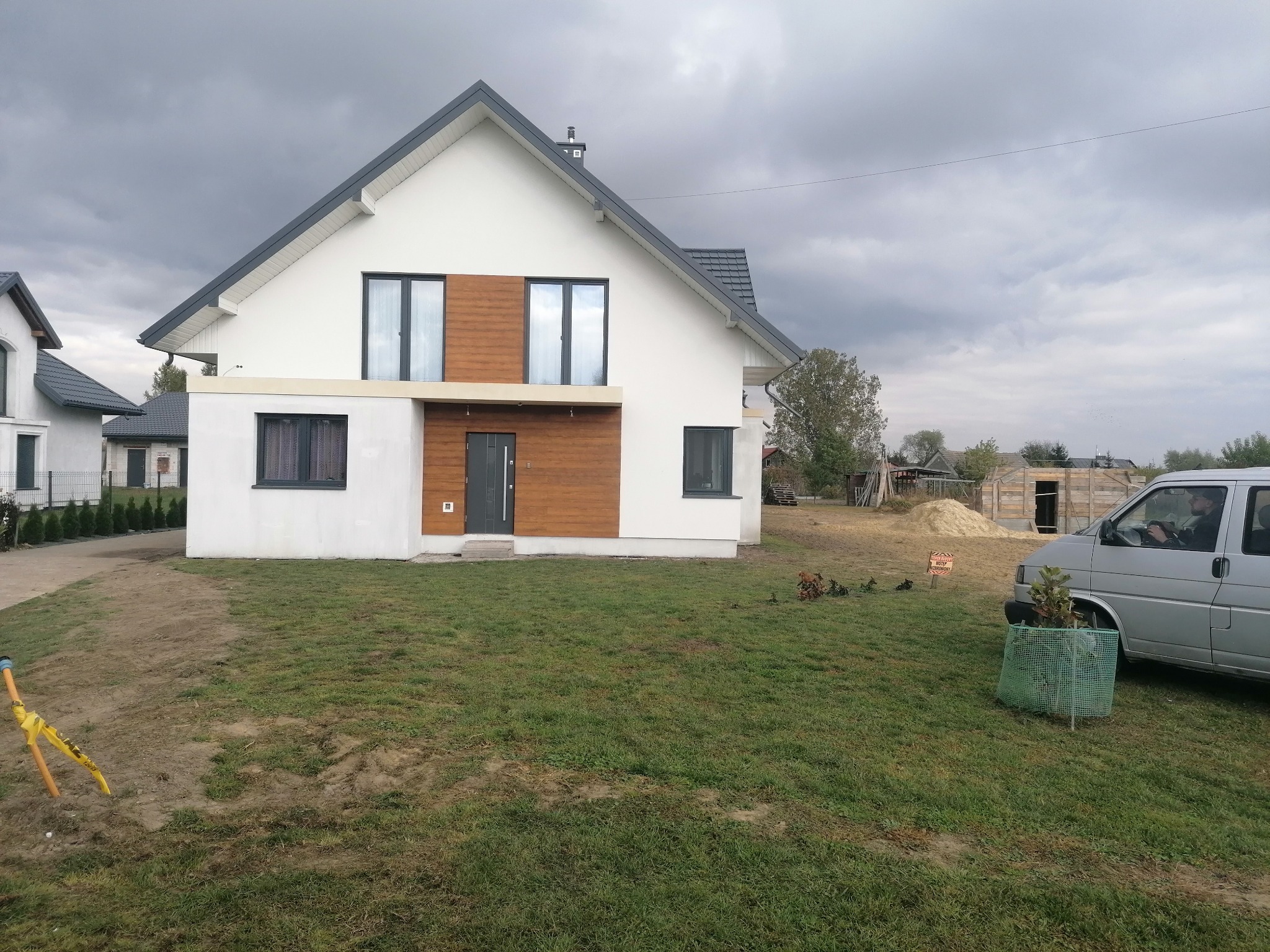 Nowoczesny dom jednorodzinny z białą elewacją i drewnianymi akcentami, szara stolarka okienna i drzwiowa, widok z zewnątrz na trawnik i zaparkowany samochód dostawczy.