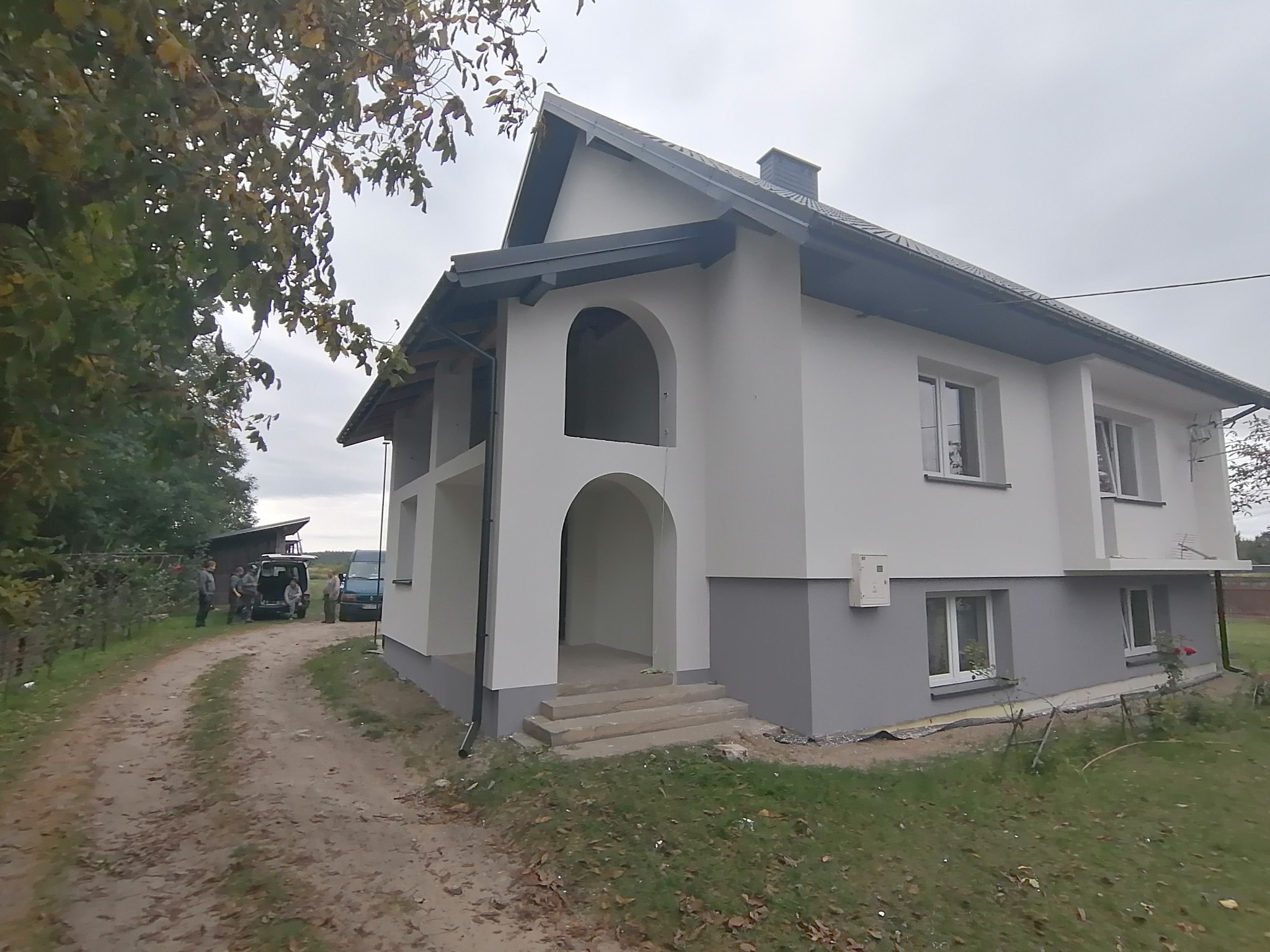 Dwukondygnacyjny dom jednorodzinny z szarą podmurówką i białymi ścianami, widoczne wykończone elewacje, z przodu wejście z łukowym portalem, ekipa remontowa przy samochodzie w tle.