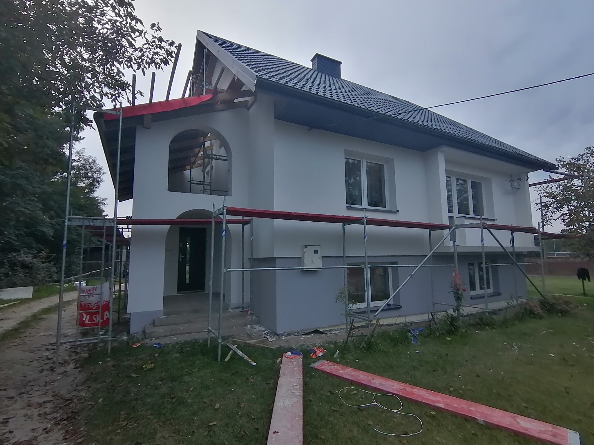 Dwukondygnacyjny dom jednorodzinny w trakcie malowania elewacji, widoczne rusztowania i częściowo pomalowana dolna partia budynku na szaro.