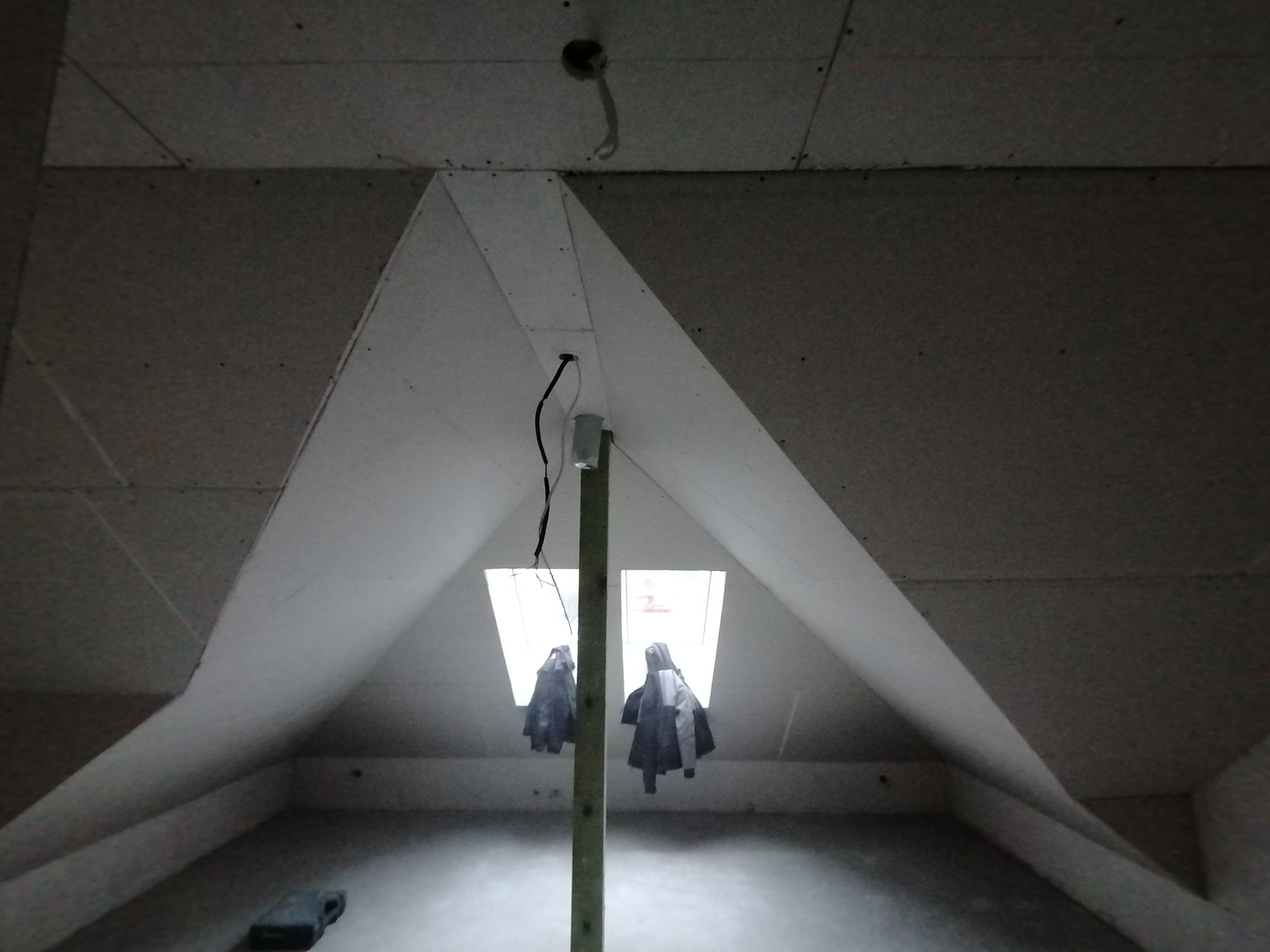 Poddasze w trakcie prac wykończeniowych: widoczne płyty kartonowo-gipsowe na skosach, drewniany słup podtrzymujący konstrukcję, dwa okna dachowe i wiszące kurtki. Na suficie widoczny otwór na kabel.