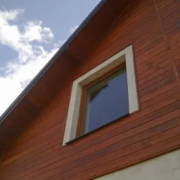 Fragment elewacji budynku z boazerią i podbitką dachową w kolorze ciemnego drewna, okno z białą ramą, widoczne niebo z chmurami.