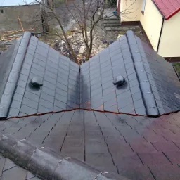 Widok z góry na nowy dach z ciemnej dachówki karpiówki z dwoma kominkami wentylacyjnymi, na tle budynków mieszkalnych i drzew bez liści.