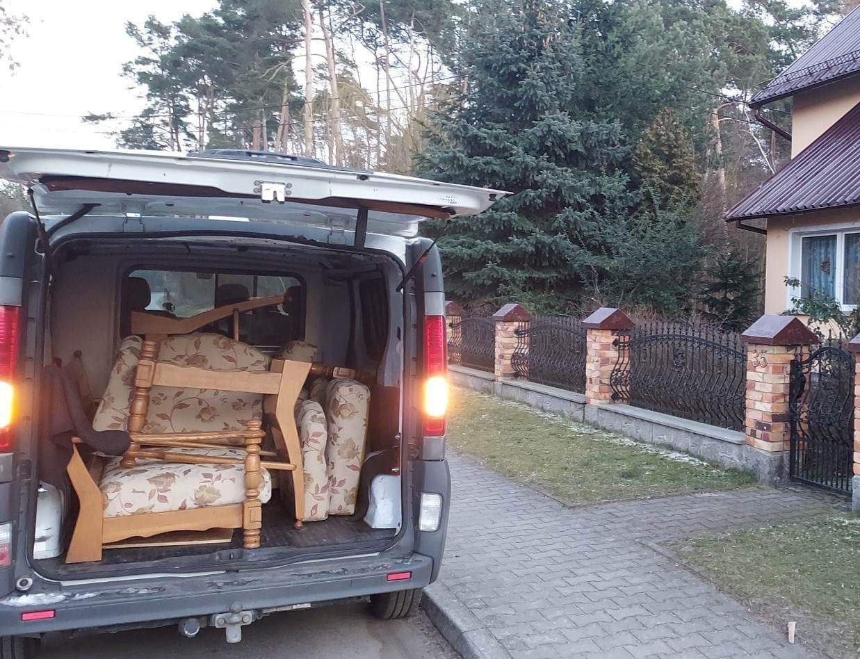 Tył szarego busa z otwartymi drzwiami, załadowanego drewnianymi fotelami z beżową tapicerką w kwiaty, na tle domu z ceglanym ogrodzeniem.