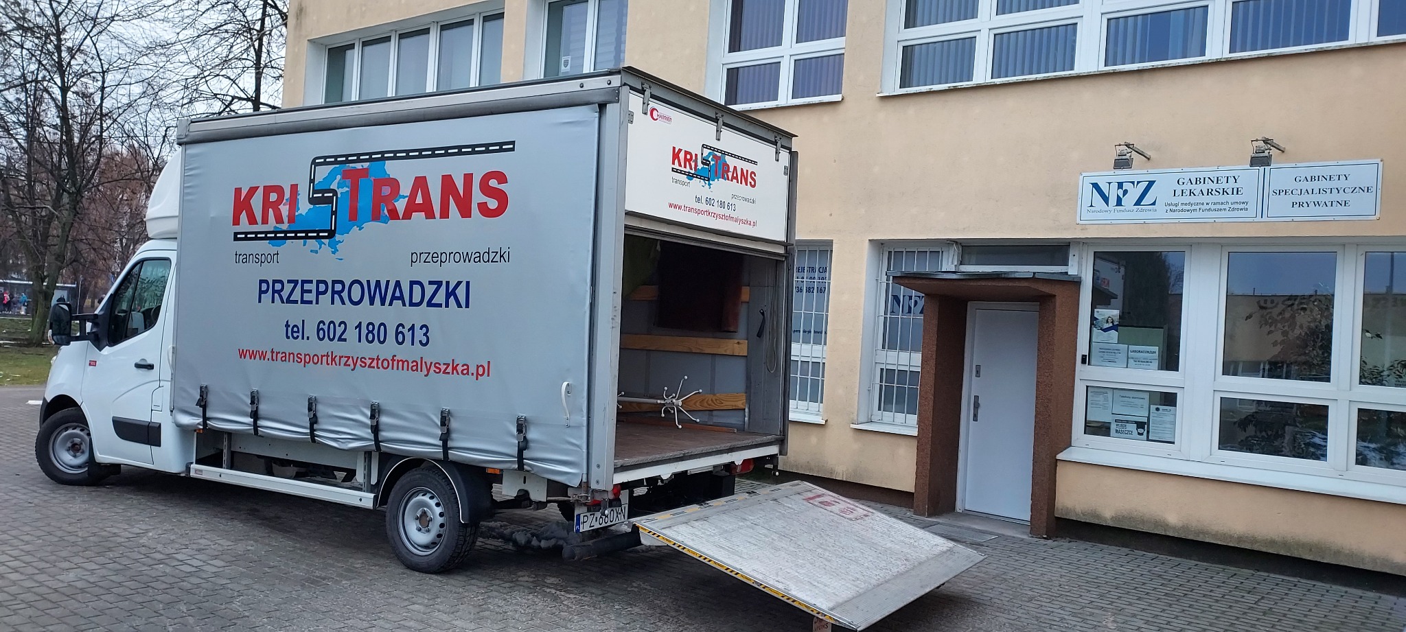 Biały bus z otwartymi drzwiami i rampą najazdową firmy KRITRANS transport przeprowadzki zaparkowany przed budynkiem z gabinetami lekarskimi NFZ i prywatnymi, wewnątrz widoczne meble i wieszak...