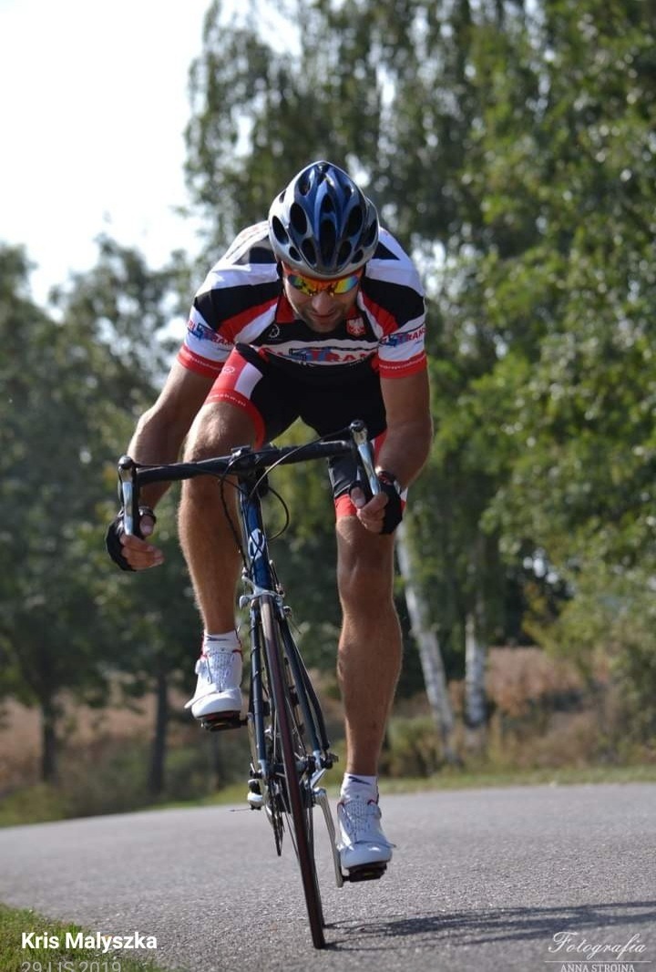 Kolarz w kasku i okularach, pochylony nad kierownicą roweru szosowego, w trakcie jazdy po asfaltowej drodze w słoneczny dzień.