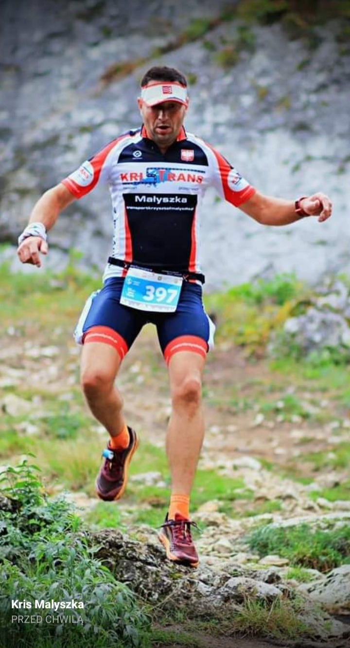 Mężczyzna w sportowym stroju z numerem 397 biegnie po kamienistej ścieżce w górach, widoczny logotyp firmy na koszulce.
