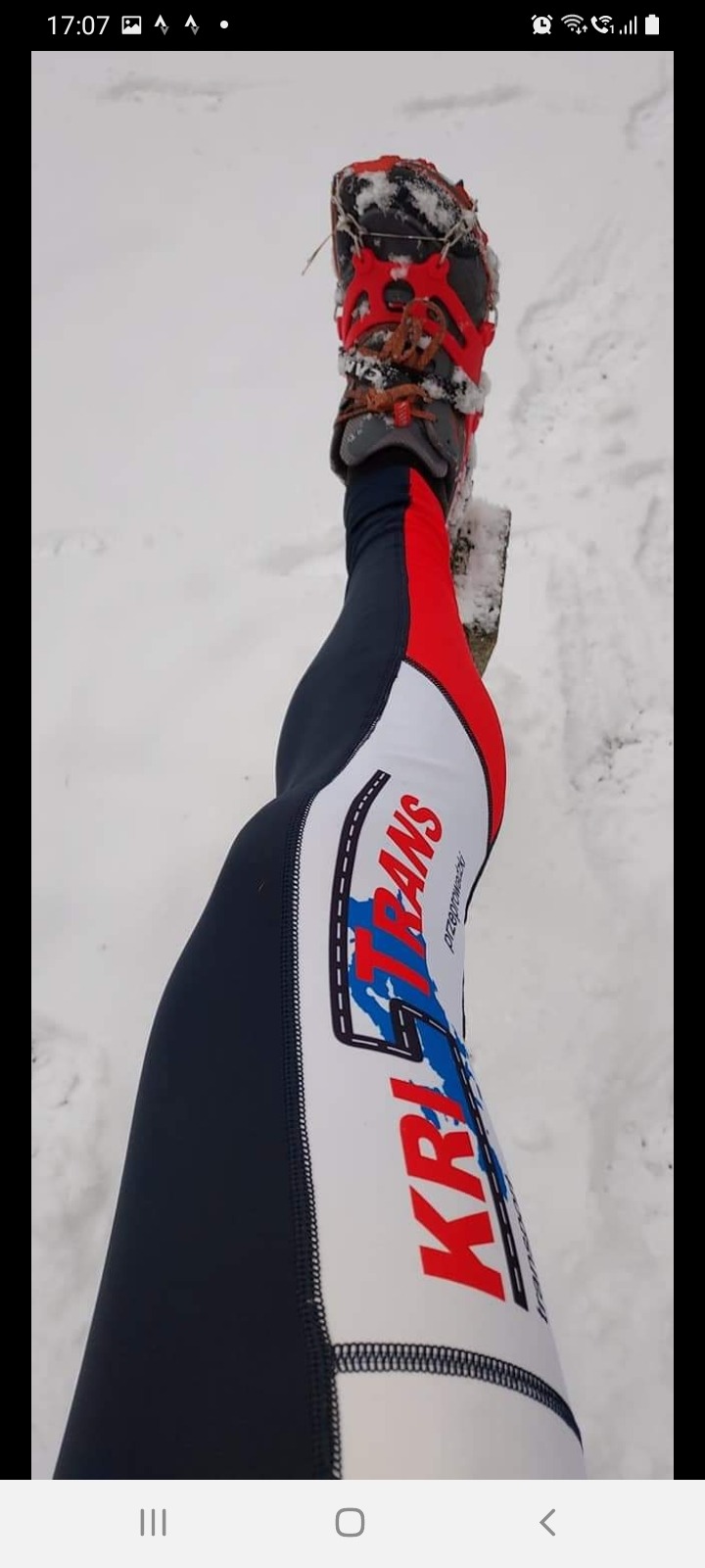 Noga w sportowych legginsach z logo firmy przeprowadzkowej i butem trekkingowym z nakładkami antypoślizgowymi, stojąca na śniegu.