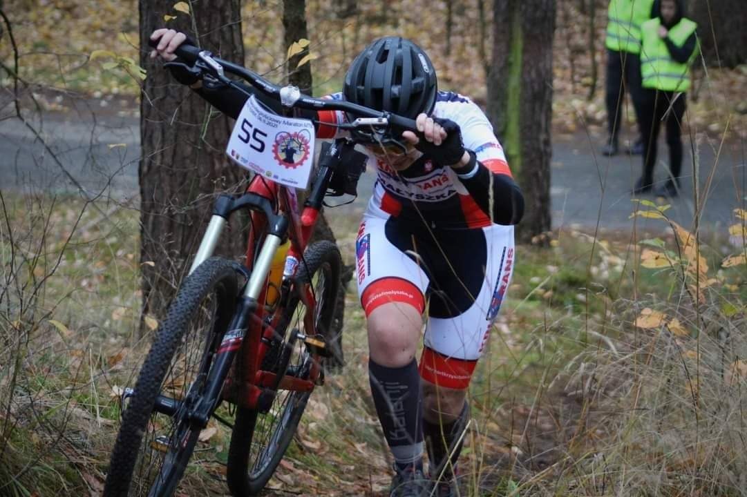 Rowerzysta w kasku prowadzi rower pod górę w lesie podczas zawodów, widoczny numer startowy 55.