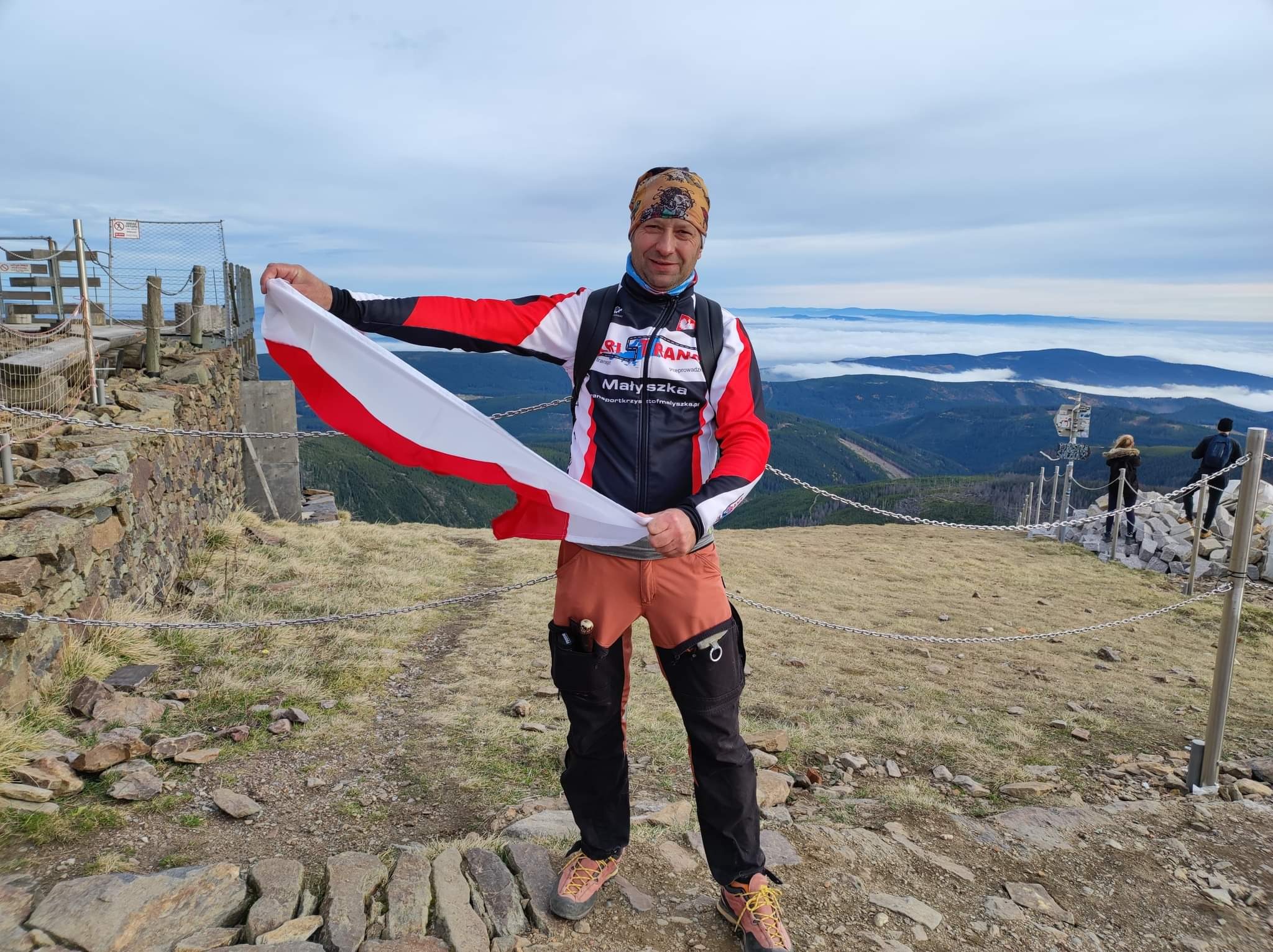 Mężczyzna na szczycie góry trzymający flagę Polski, w tle widok na góry i chmury poniżej szczytu.