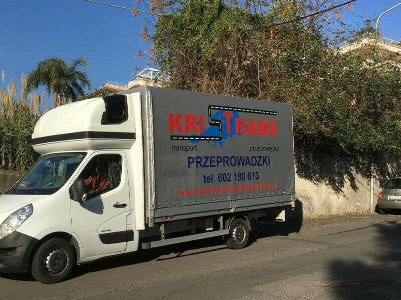 Biały bus dostawczy z szarym plandekowym nadwoziem, na którym widnieje logo firmy transportowej 'KRI-TRANS' z numerem telefonu, zaparkowany na ulicy w słoneczny dzień.