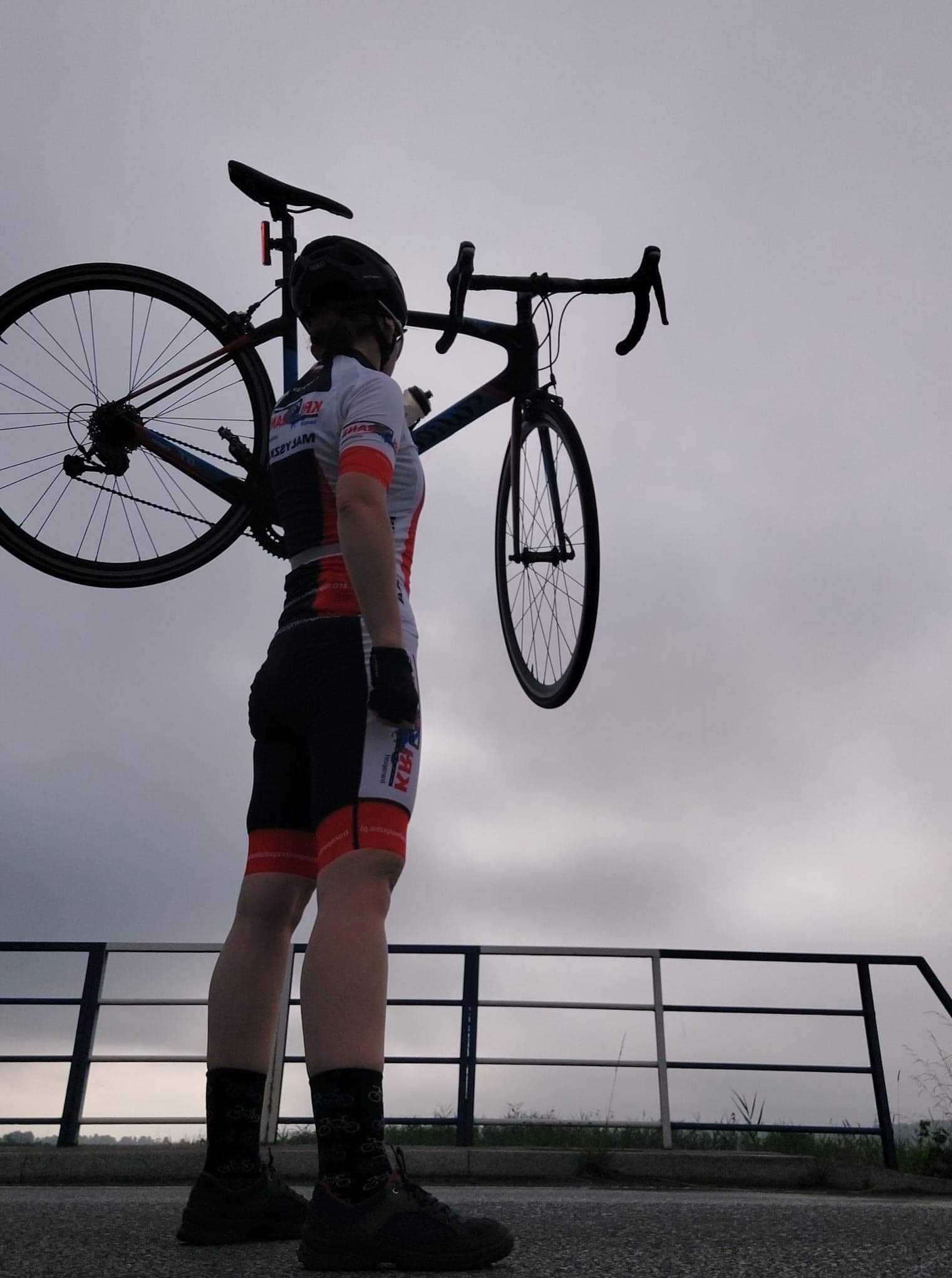 Kolarz w stroju sportowym niesie rower wyścigowy na ramieniu, patrząc w dal na pochmurne niebo