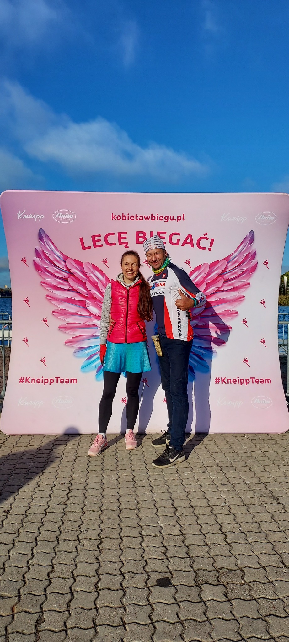 Dwoje uśmiechniętych biegaczy pozuje na tle ścianki reklamowej z napisem 'Lecę biegać!' i logotypami sponsorów Kneipp i Anita.