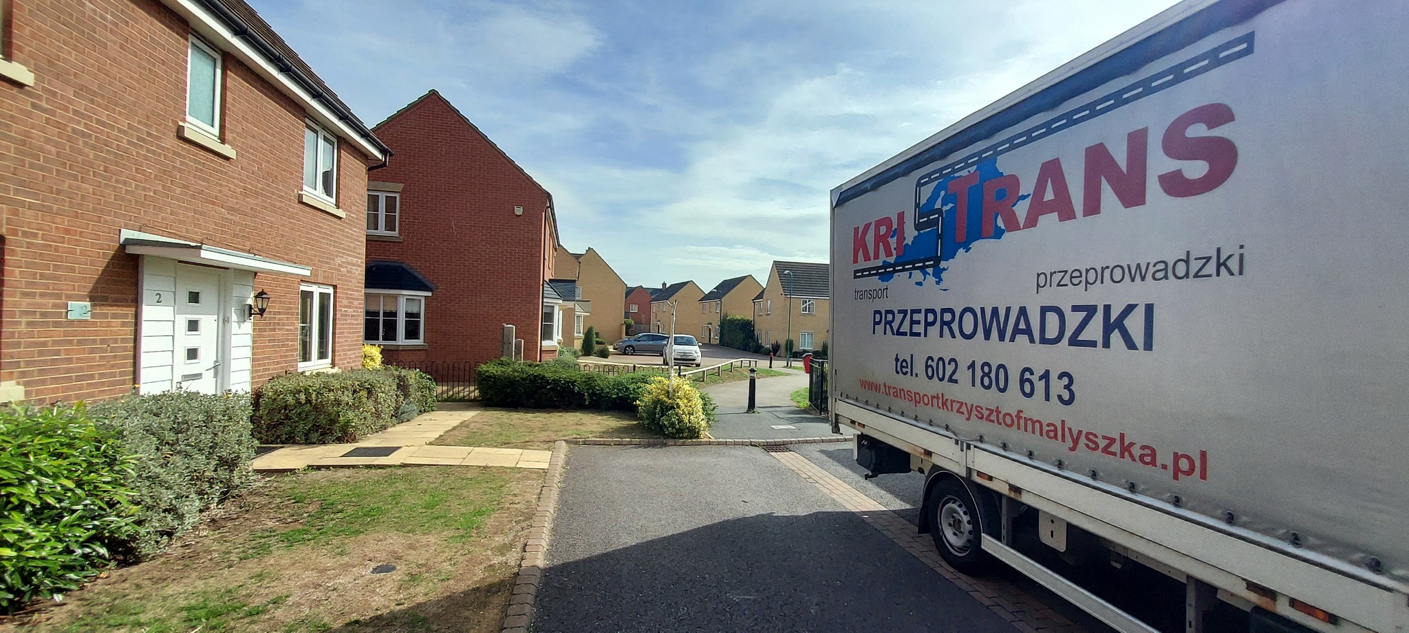 Ciężarówka z napisem 'KRI TRANS transport przeprowadzki' zaparkowana przed domami jednorodzinnymi na osiedlu.
