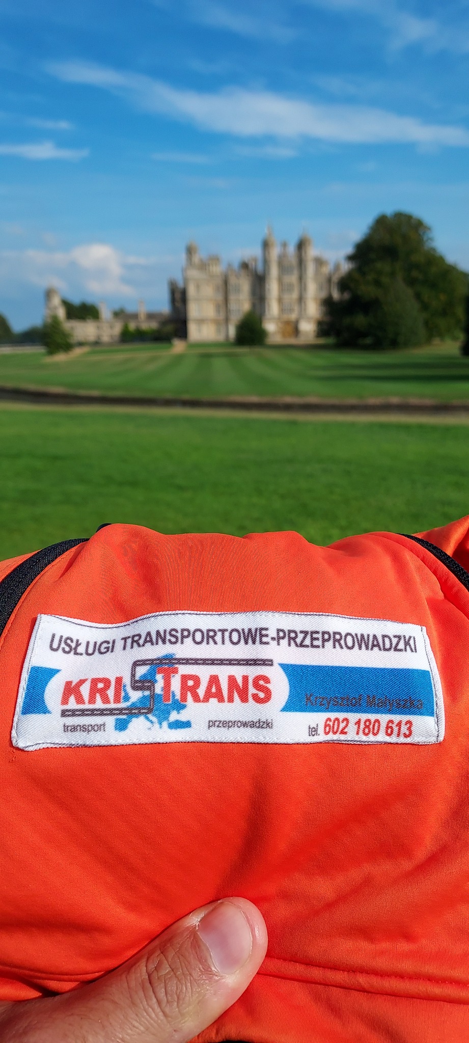 Pomarańczowa torba z logo firmy transportowej KRISTRANS na tle rozległego, zielonego trawnika i oddalonego zamku w stylu angielskim.