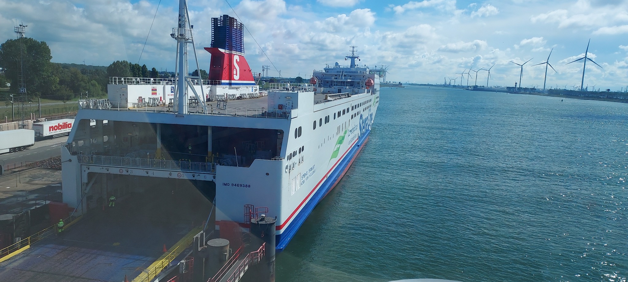 Duży prom pasażerski Stena Line zacumowany w porcie, z otwartą rampą załadunkową, w tle widać turbiny wiatrowe na brzegu.