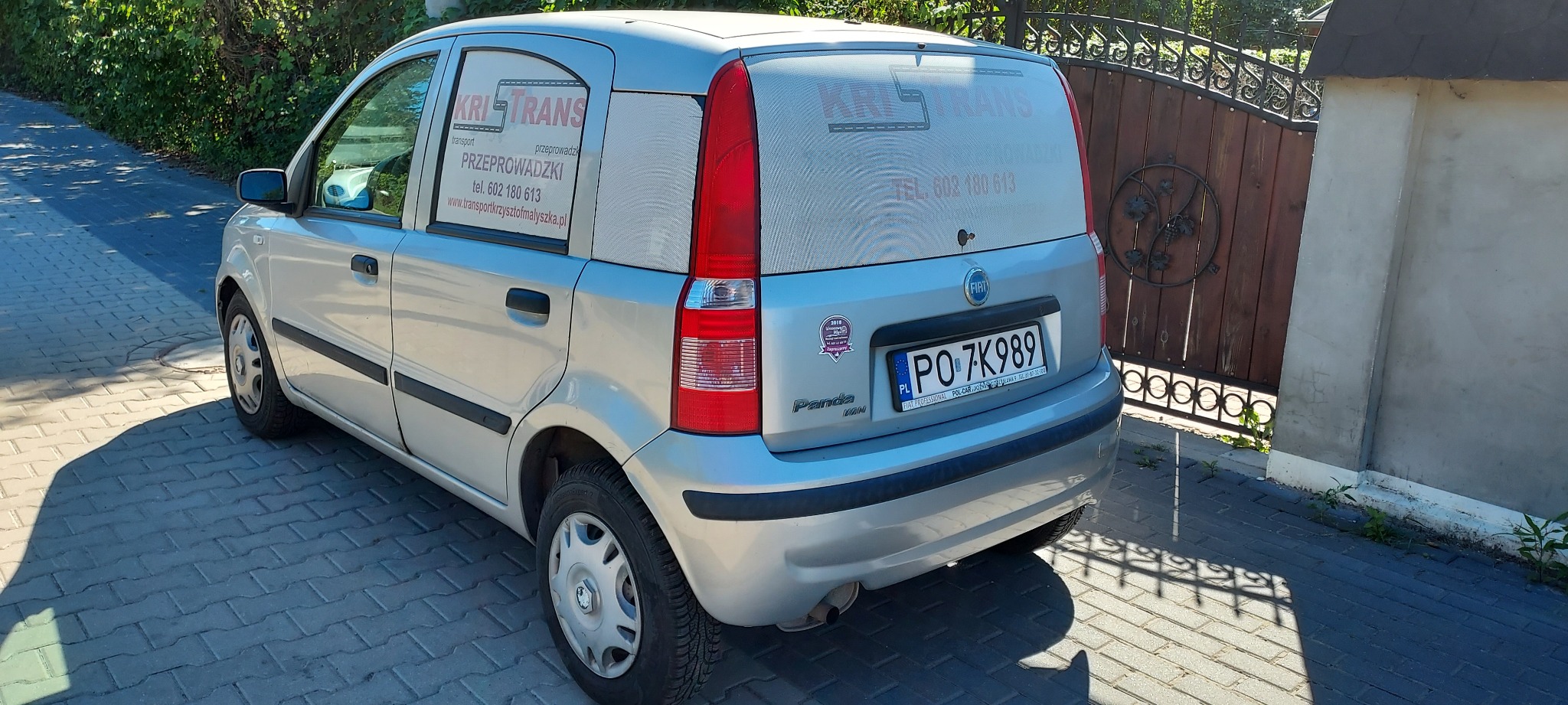 Srebrny samochód dostawczy Fiat Panda Van z reklamą firmy transportowej KRI-TRANS na szybach, zaparkowany na brukowanej drodze przed drewnianą bramą.