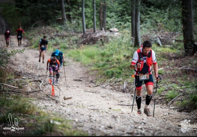 Grupa biegaczy górskich pokonuje strome podejście leśną ścieżką, używając kijków trekkingowych, z widocznymi numerami startowymi i logotypami organizatorów na odzieży i w tle drzewa.