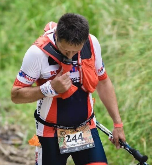 Mężczyzna w sportowym stroju z numerem 244 przypiętym do pasa, trzymający kijki trekkingowe, prawdopodobnie podczas biegu górskiego.