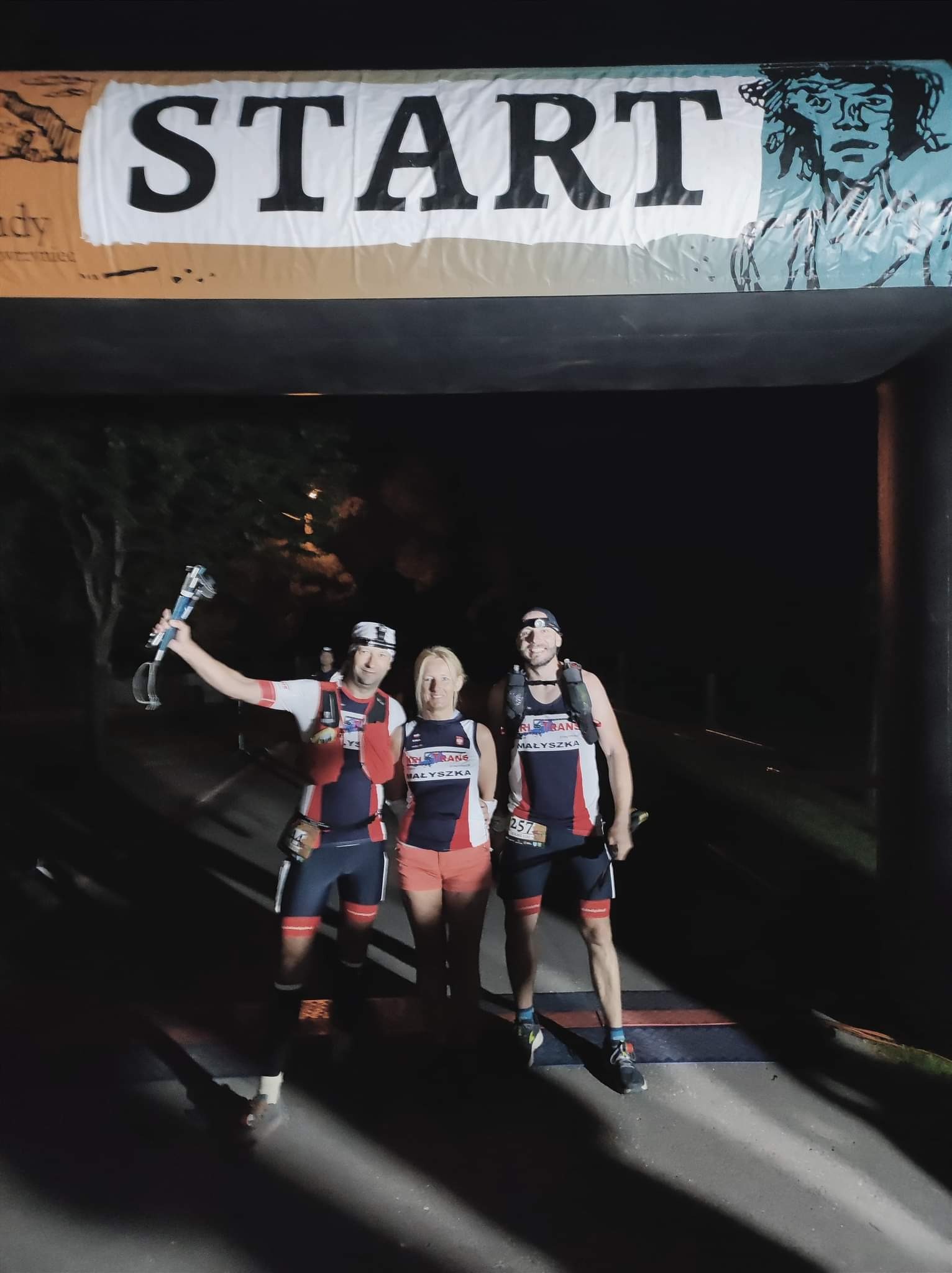Trójka uśmiechniętych biegaczy w sportowych strojach, stojąca pod banerem z napisem 'START' w nocnych warunkach oświetleniowych.