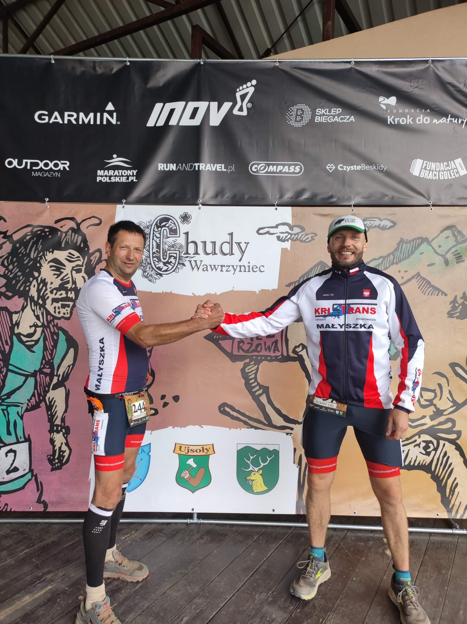 Dwóch biegaczy w strojach sportowych, z numerami startowymi, ściska sobie dłonie na tle baneru sponsorskiego po zawodach biegowych Chudy Wawrzyniec. Jeden ma na sobie bluzę z napisem 'KRI-TRANS...