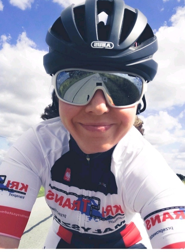 Uśmiechnięta kobieta w kasku rowerowym i okularach przeciwsłonecznych, w których odbija się droga, ubrana w koszulkę z logotypem firmy transportowej.