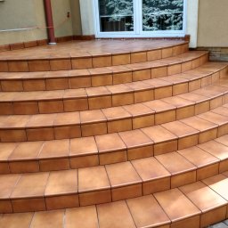 Piotr Socha - Zewnętrzne schody wejściowe do domu wyłożone brązowymi płytkami ceramicznymi, widoczne z dołu, z oknem odbijającym drzewa w tle.