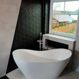 Nowoczesna łazienka z wolnostojącą białą wanną na tle ściany z czarną, geometryczną mozaiką i dużym oknem z widokiem na ogród.