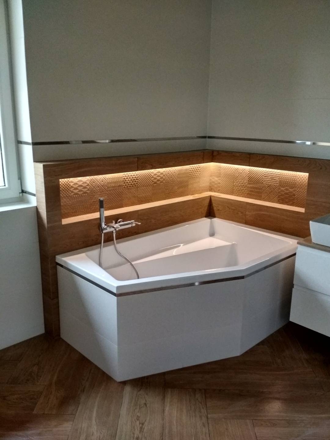 Nowoczesna narożna wanna z chromowaną armaturą, obudowana płytkami imitującymi drewno z subtelnym podświetleniem LED, na podłodze parkiet ułożony w jodełkę.