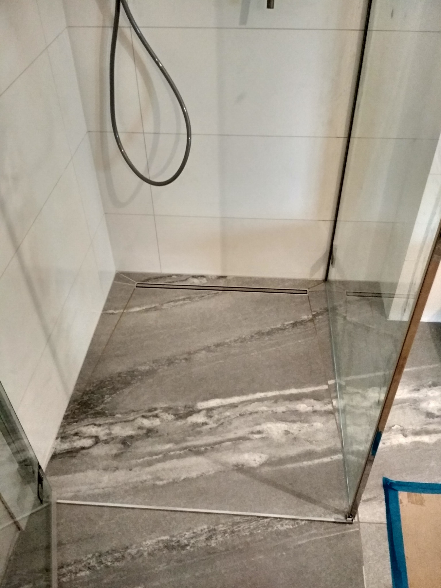 Nowoczesna kabina prysznicowa walk-in z odpływem liniowym wkomponowanym w marmurowe płytki podłogowe, widoczna szklana tafla z chromowanymi okuciami.