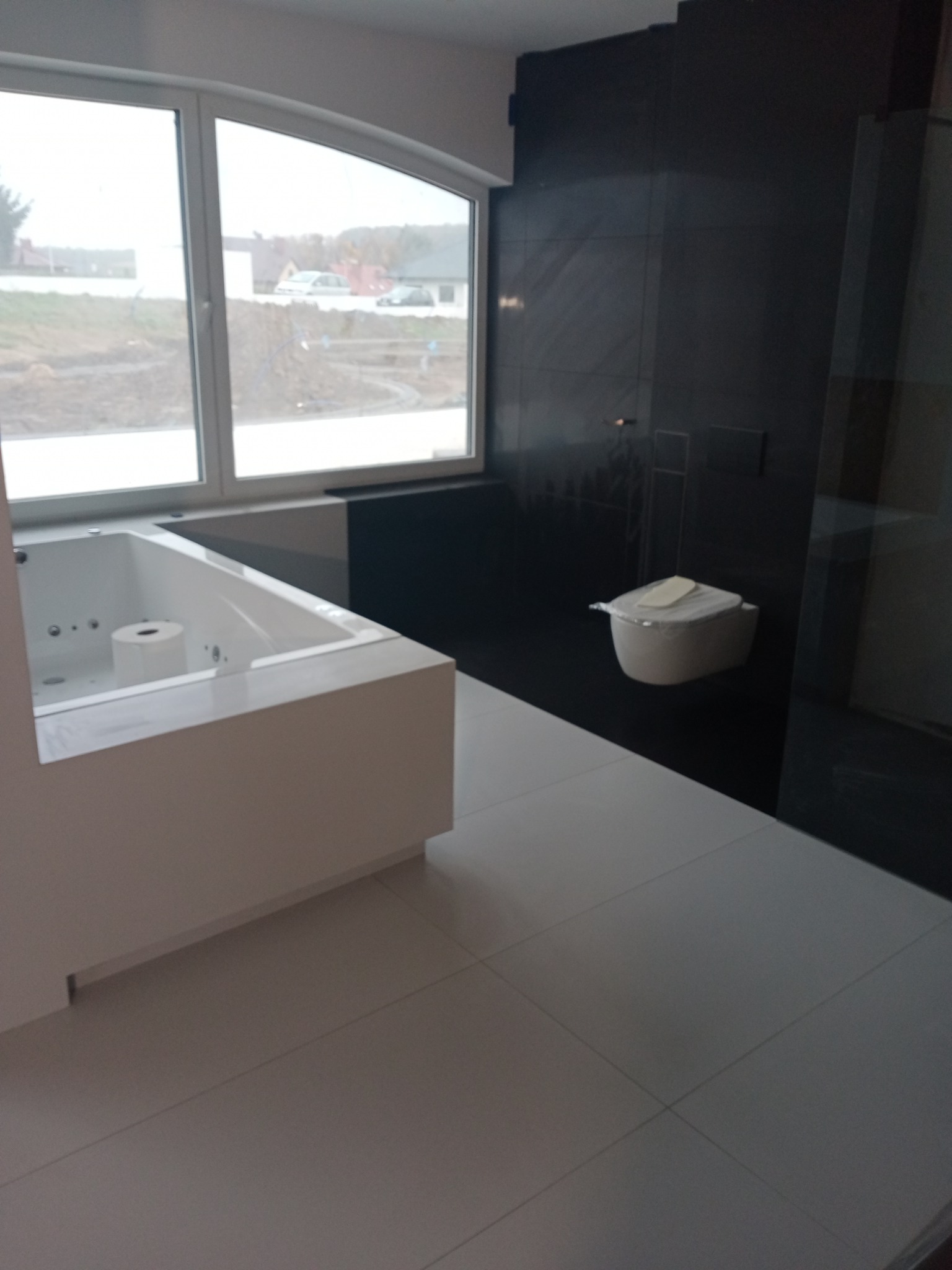 Minimalistyczna łazienka z białą wanną z hydromasażem, toaletą w folii ochronnej i czarnymi płytkami ściennymi, widok przez okno na krajobraz.