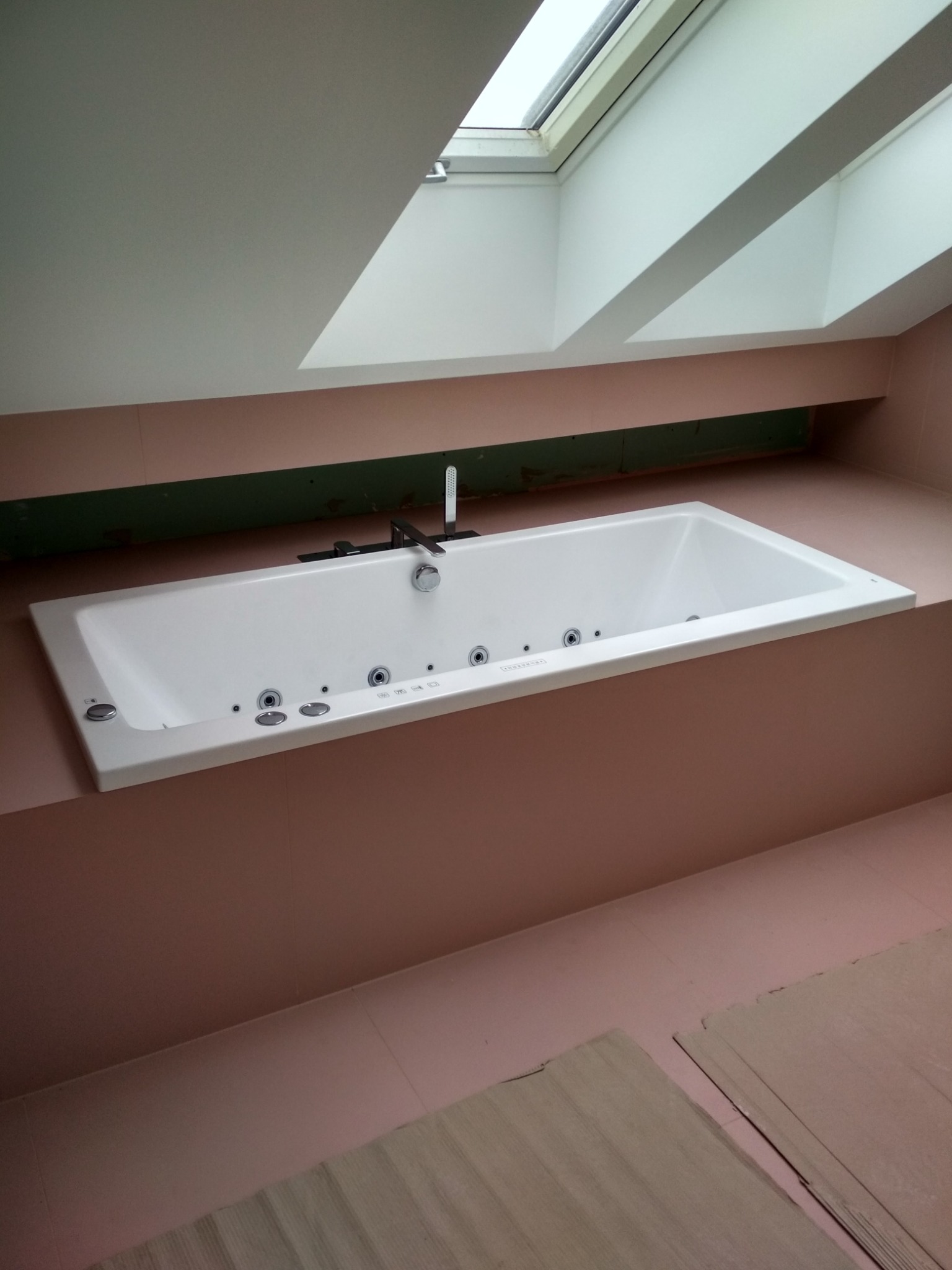Biała wanna z hydromasażem zabudowana w różowej wnęce pod oknem dachowym, widoczne fragmenty niezakończonych prac wykończeniowych na podłodze.