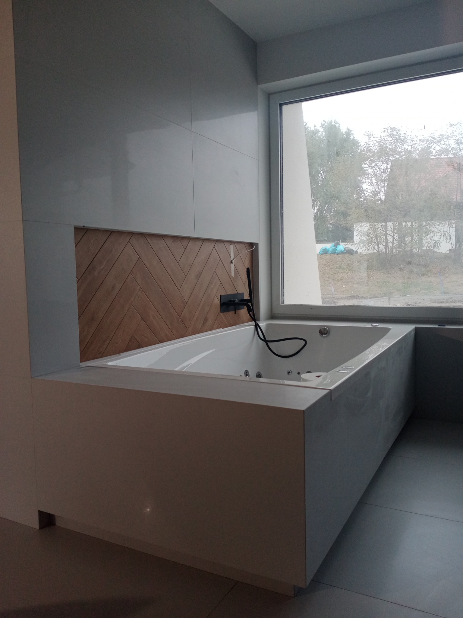 Nowoczesna wanna z hydromasażem, obudowana białą zabudową, z drewnianym panelem dekoracyjnym w jodełkę na ścianie i dużym oknem z widokiem na zewnątrz.