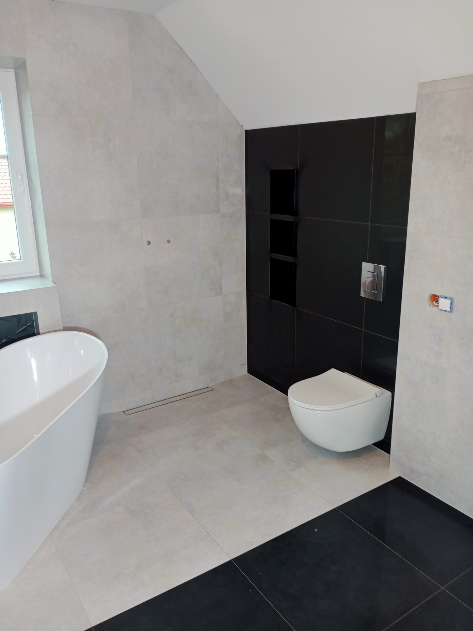 Nowoczesna łazienka z wolnostojącą wanną, czarnymi płytkami w strefie WC z wnękami i szarą ceramiką na podłodze oraz ścianach, widoczny odpływ liniowy.