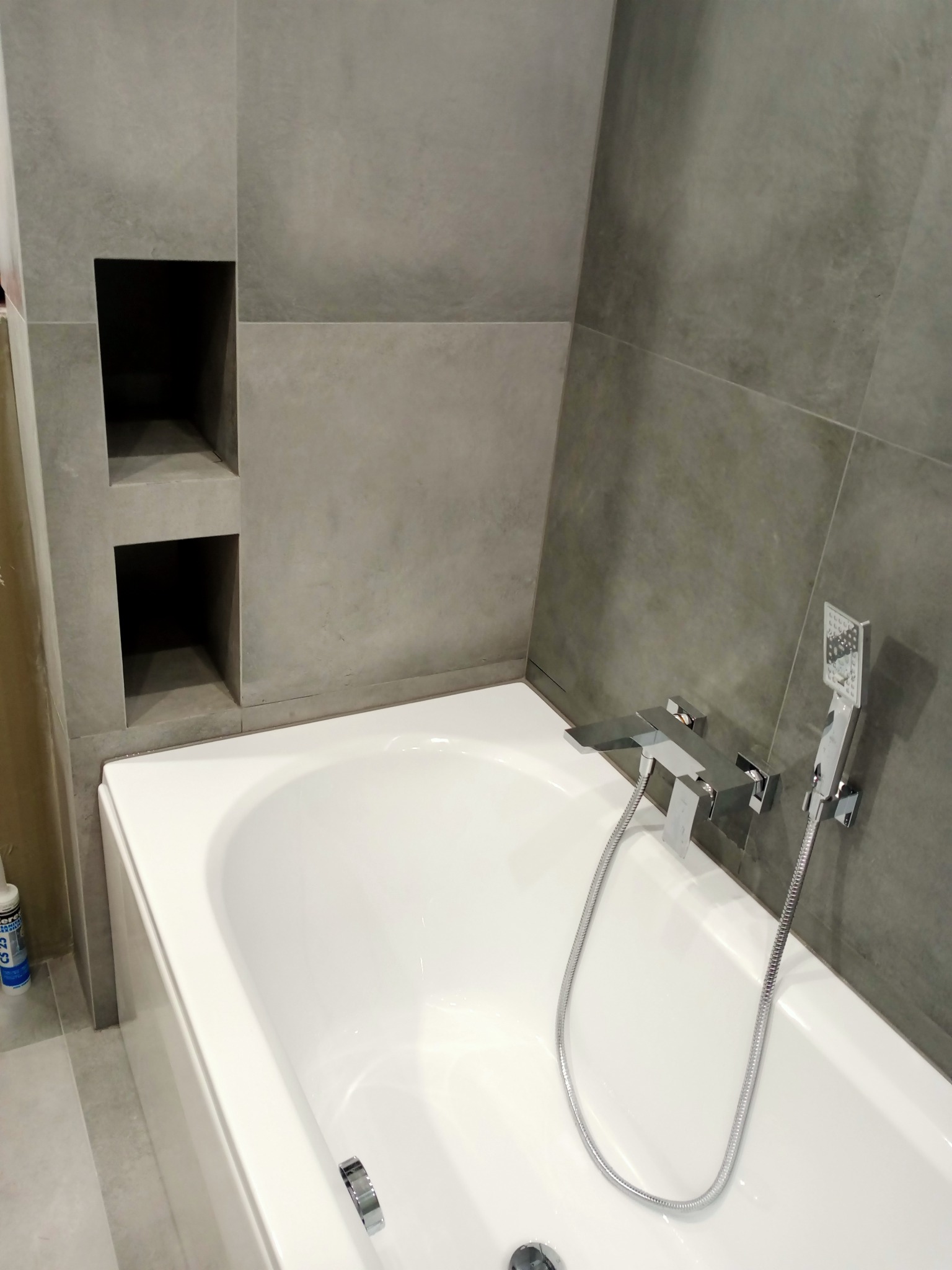 Biała wanna z chromowaną armaturą i prysznicem na tle ściany wyłożonej szarymi płytkami z wnękami.