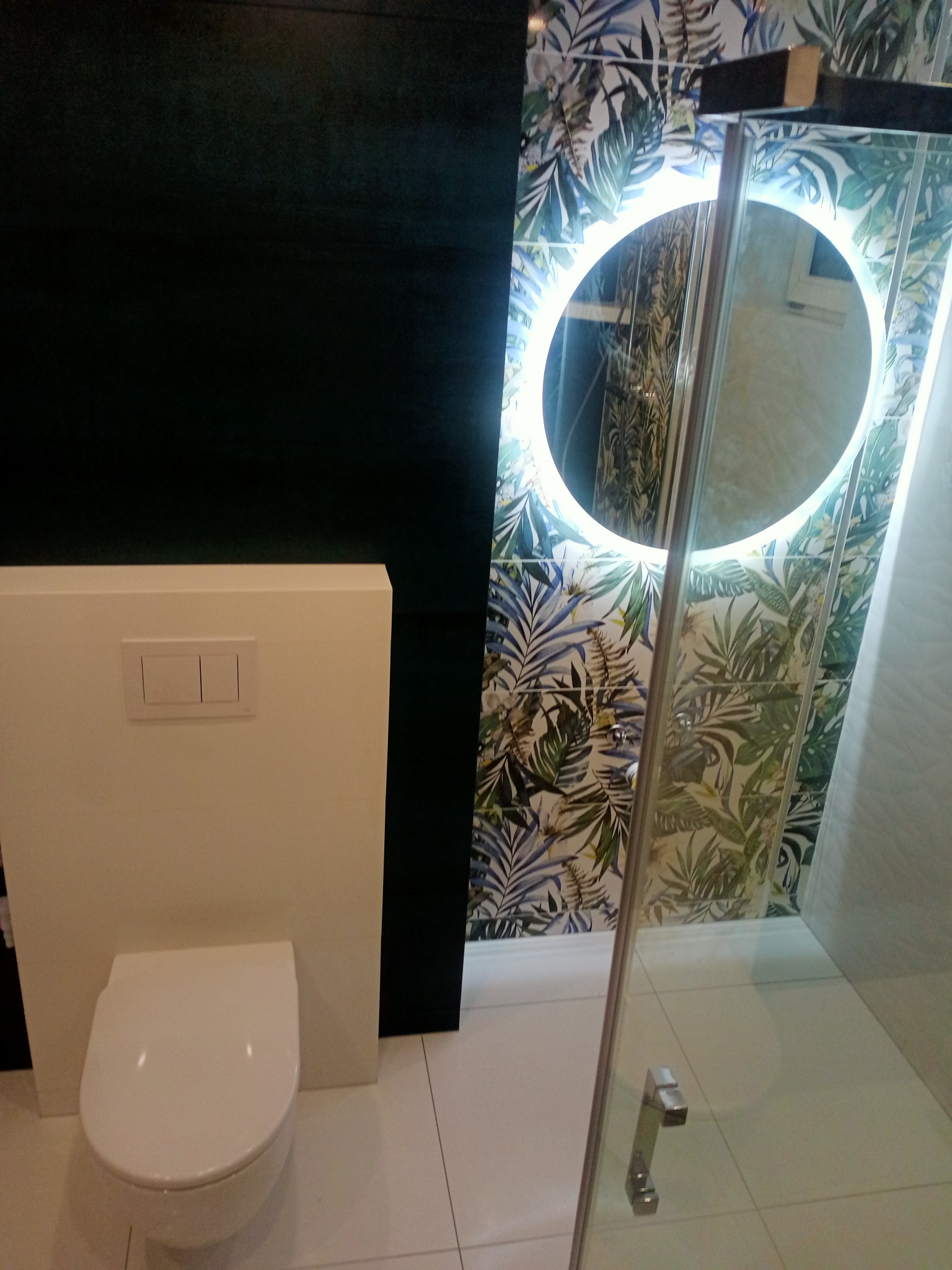 Nowoczesna łazienka z podwieszaną toaletą, czarną ścianą, kabiną prysznicową i okrągłym lustrem z podświetleniem LED na tle tropikalnych płytek.